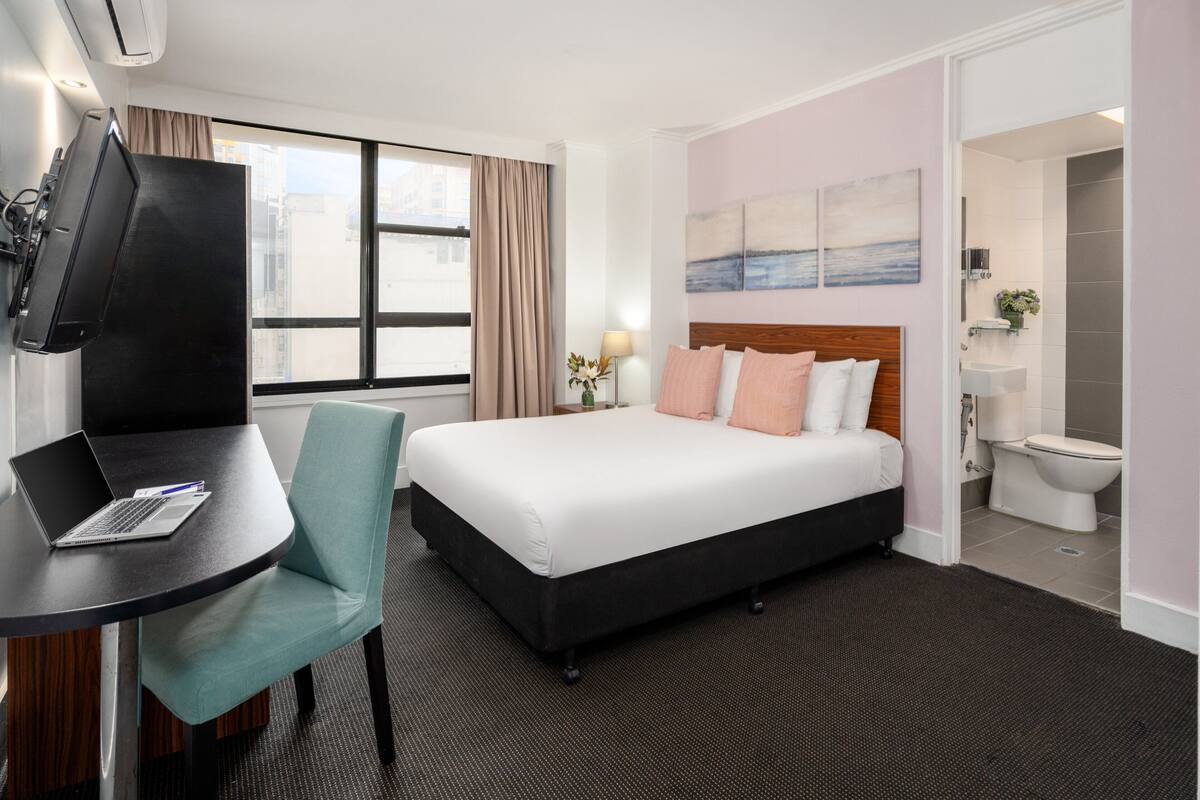 A hotel room with a bed, a desk with a laptop, a TV, and a view of the city through a window.