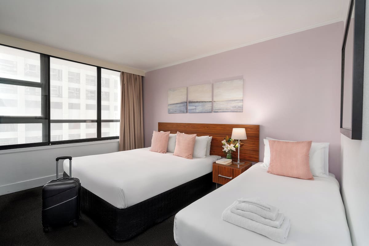 A hotel room with two beds, a suitcase, a nightstand with a lamp, and a window with curtains.
