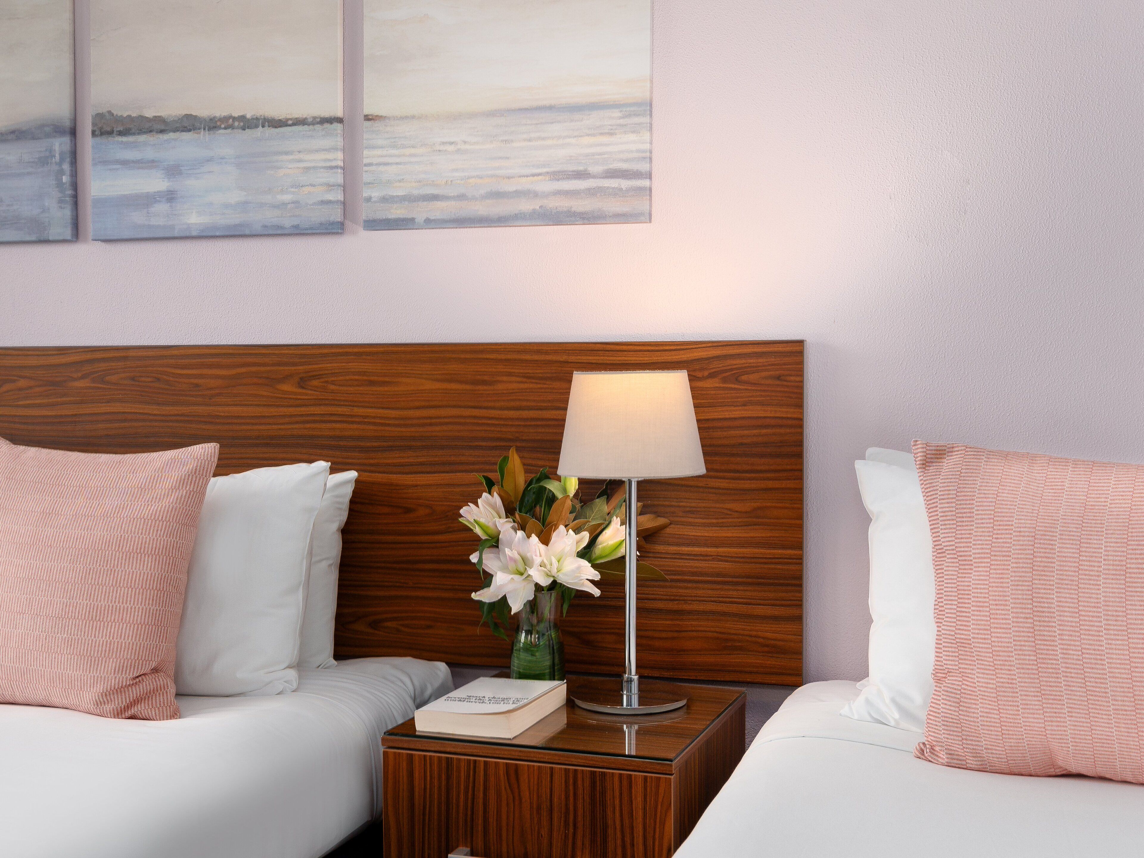 A hotel room with two beds, a wooden headboard, a nightstand with a lamp and a book, and a painting on the wall.