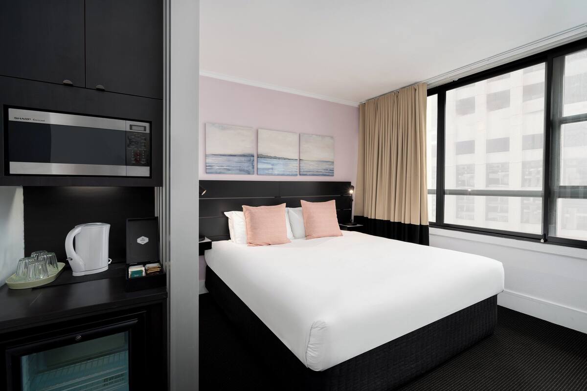 A hotel room with a bed, a minibar, a microwave, and a large window with curtains.