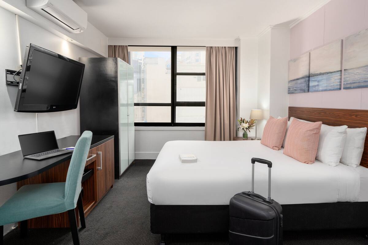 A hotel room with a bed, a desk with a computer, a TV, and a window with curtains.