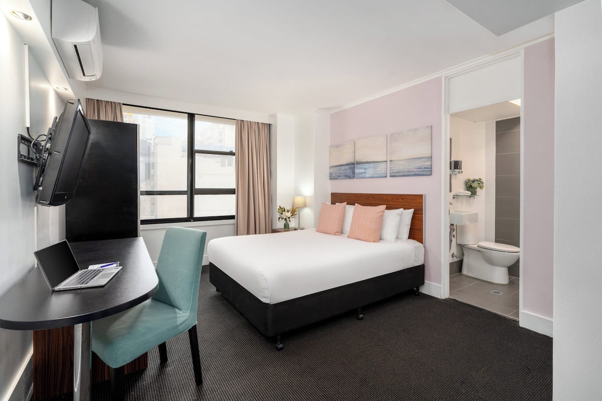 A hotel room with a bed, a desk with a laptop, a chair, a TV, and a bathroom.