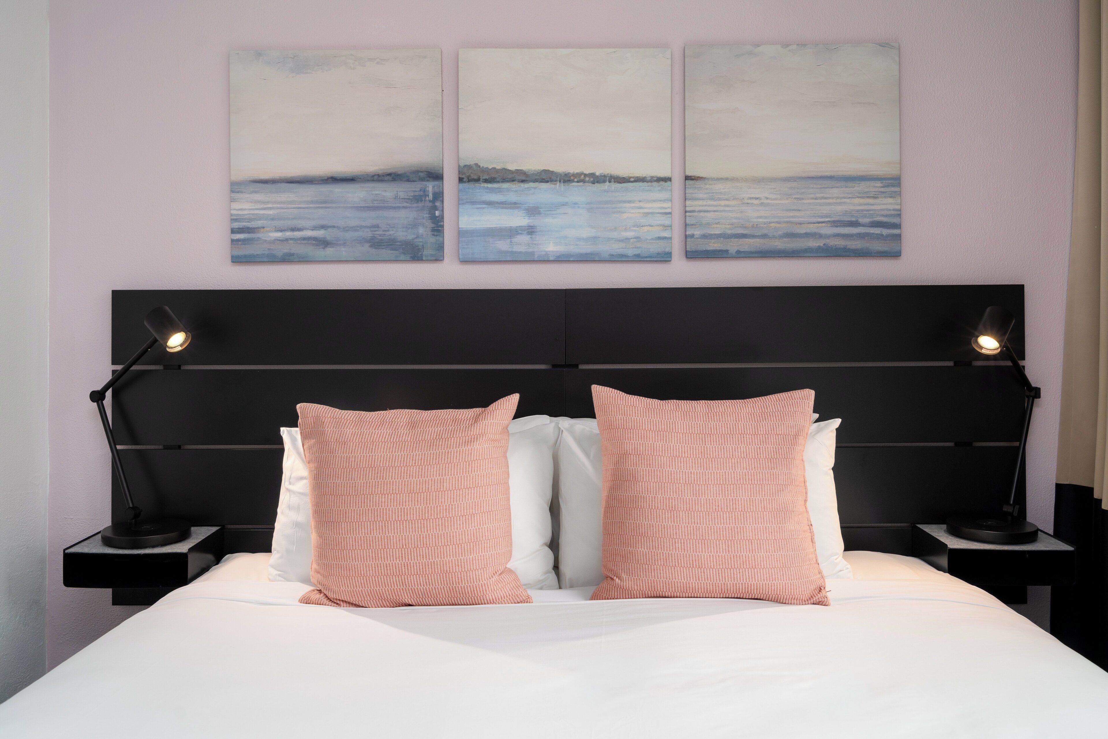 Una cama bien hecha con dos almohadas rosas y una blanca, flanqueada por dos mesitas de noche con lámparas, frente a una pared con tres cuadros abstractos.