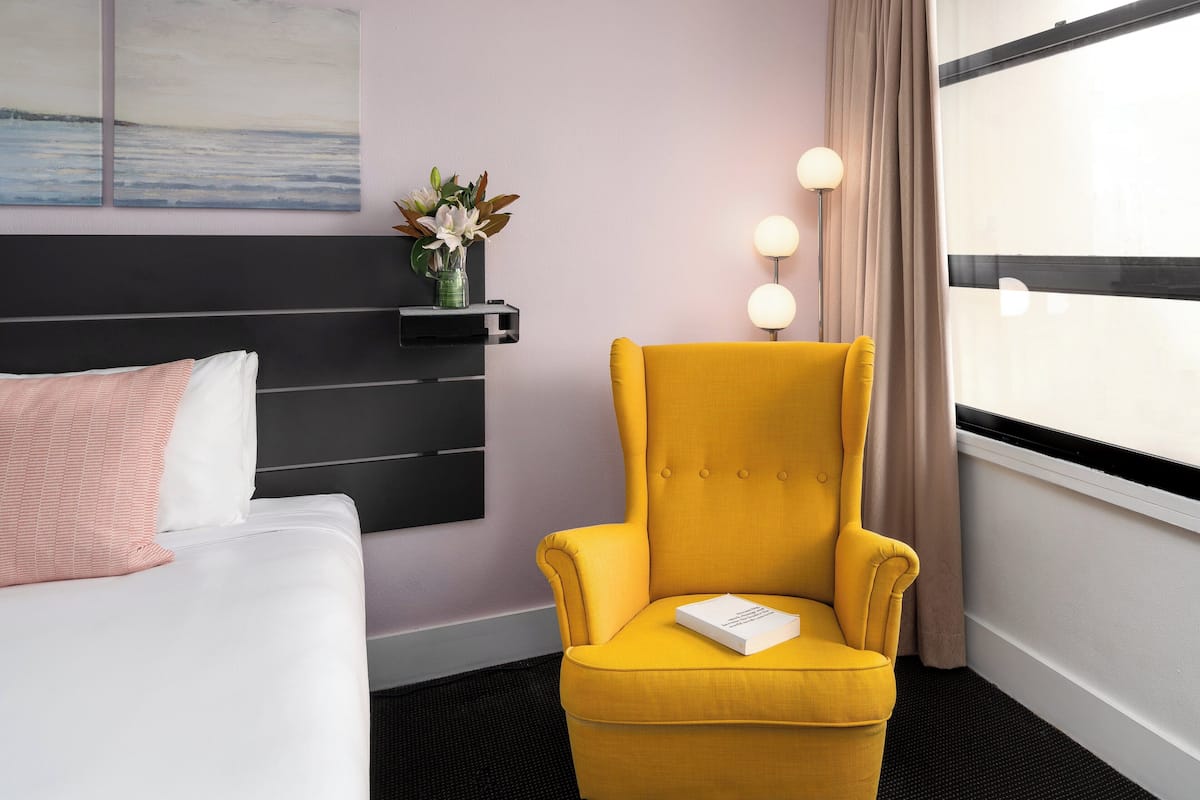 A hotel room with a bed, a yellow armchair, a bedside table with a vase of flowers, and a large window with curtains.