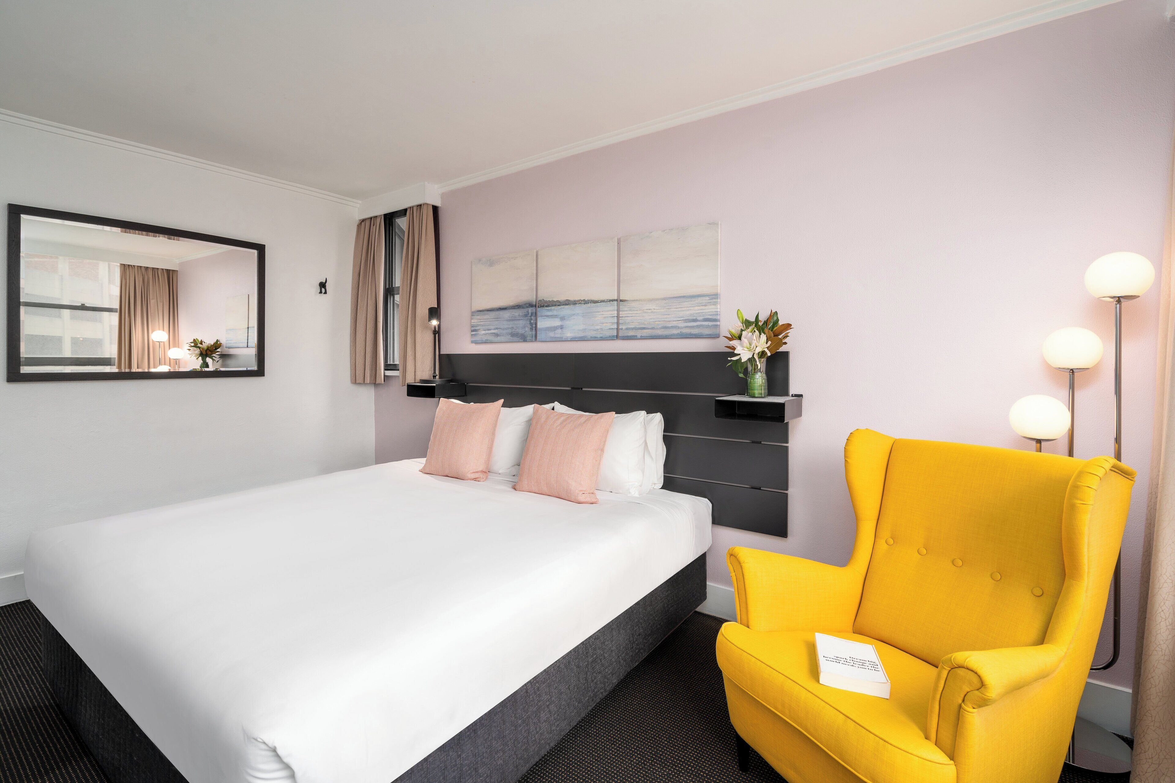 A hotel room with a large bed, a yellow armchair, a bedside table with a lamp, a mirror, and a wall-mounted artwork.