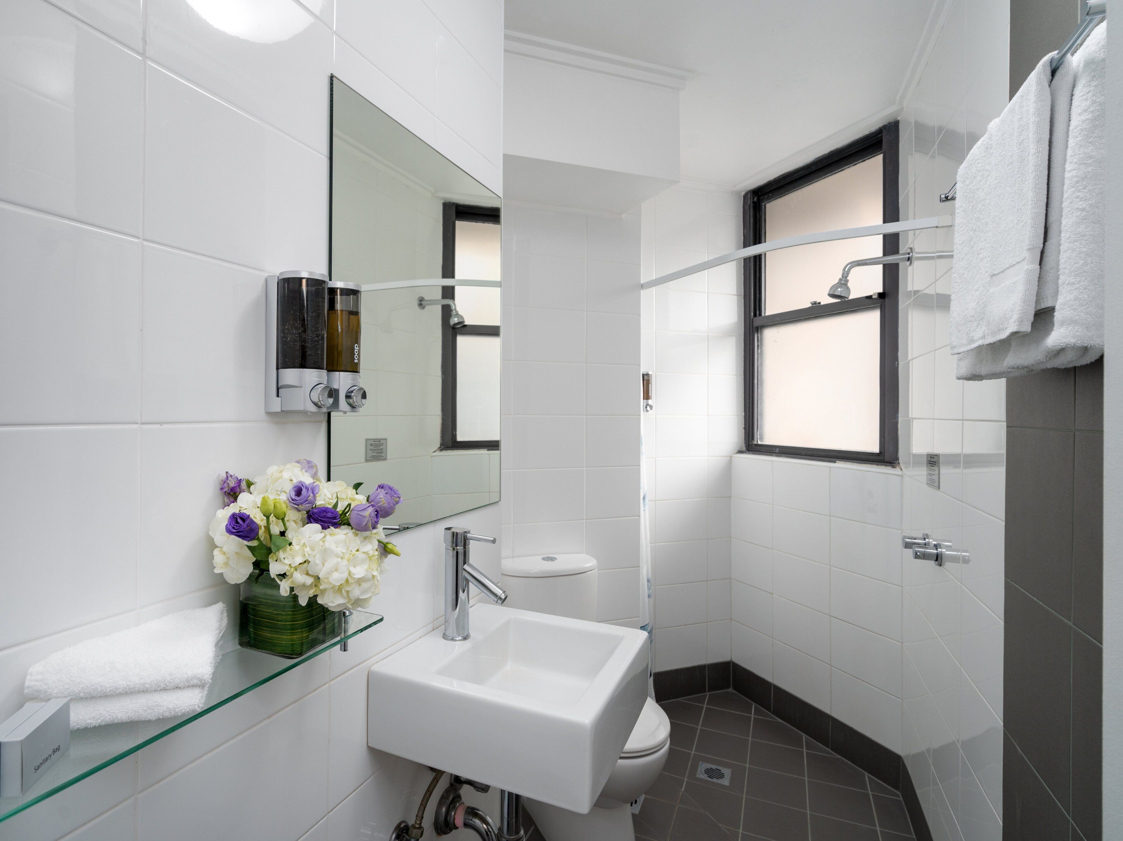 A modern bathroom with a glass shelf holding a vase of flowers, a white sink, a mirror, and a shower area.