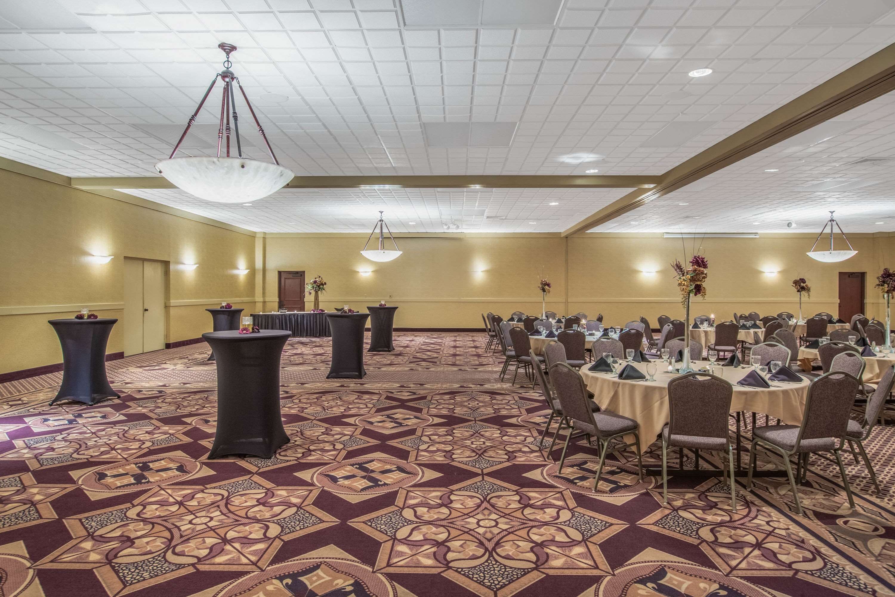 Une salle de banquet avec des tables rondes dressées pour le repas, une moquette à motifs et des luminaires au plafond.