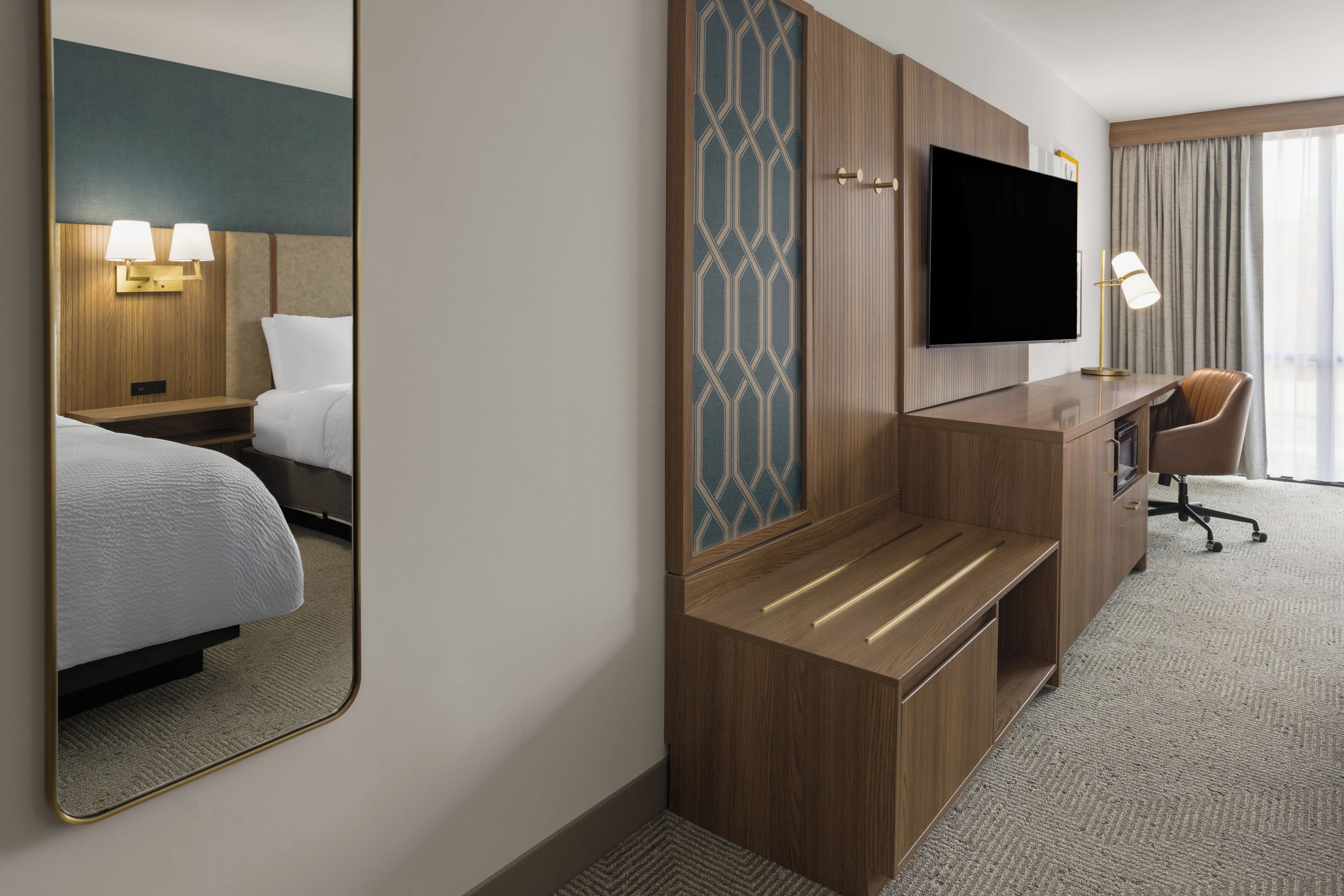 A hotel room with a bed, a desk, a chair, a flat-screen TV, and a mirror.
