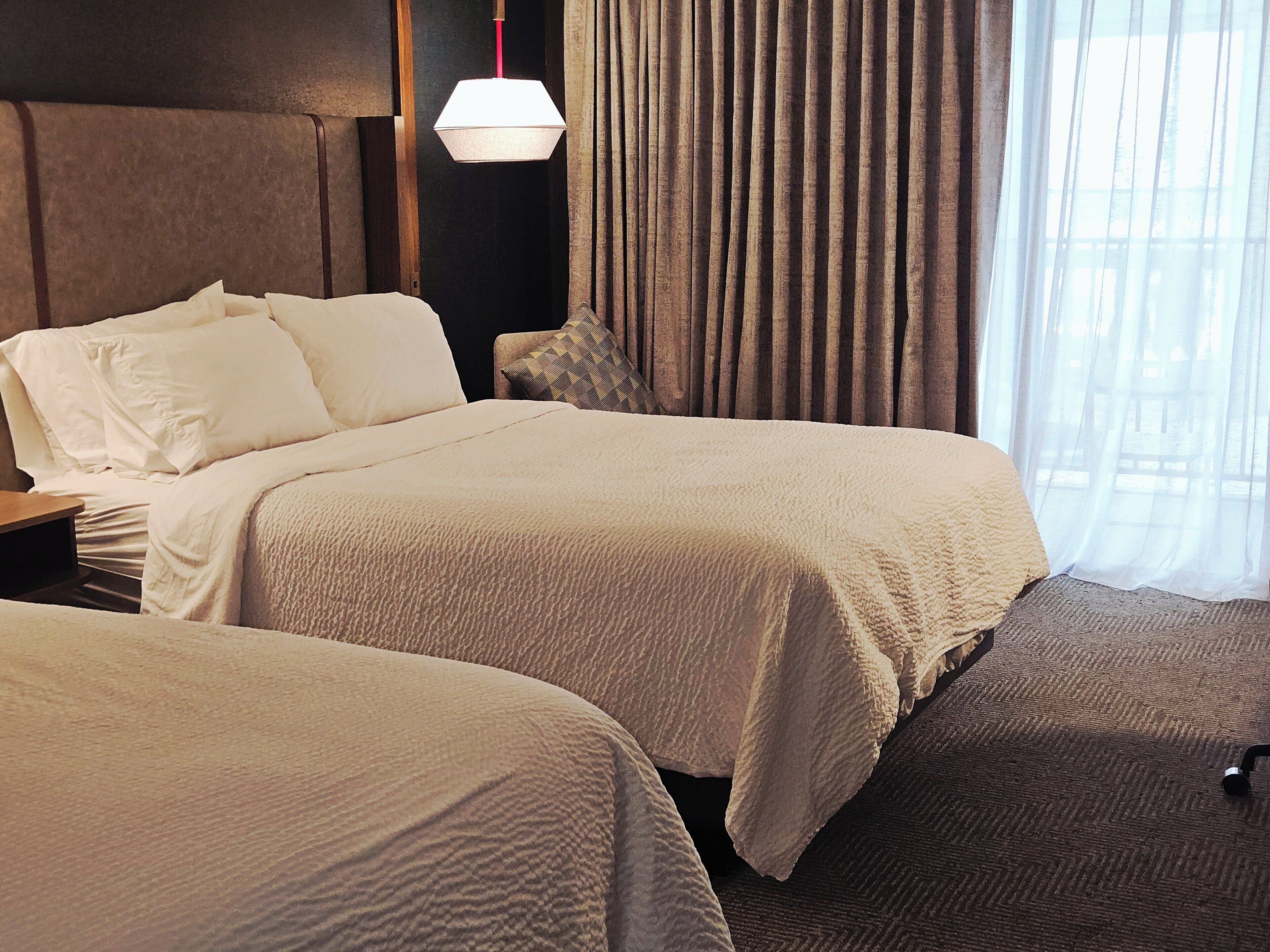 Habitación de hotel con dos camas, una mesita de noche, una lámpara y vistas al exterior.