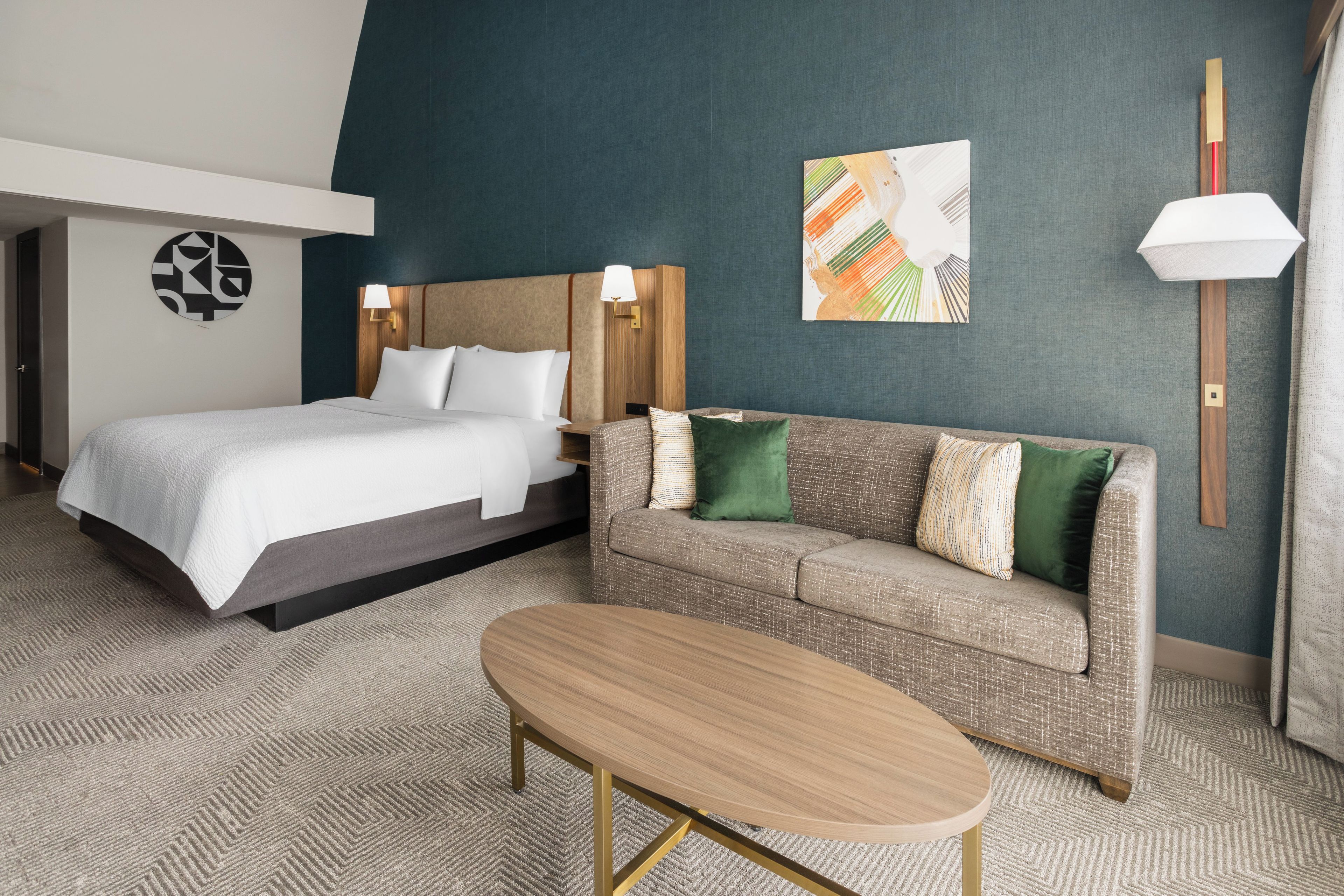 A hotel room with a bed, a sofa, a table, and a wall with a clock and artwork.