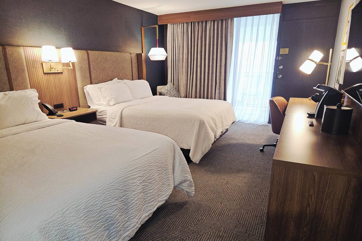 Ein Hotelzimmer mit zwei Betten, einem Schreibtisch, einem Stuhl, einer Lampe und einem fensterbepflanzten Fenster.