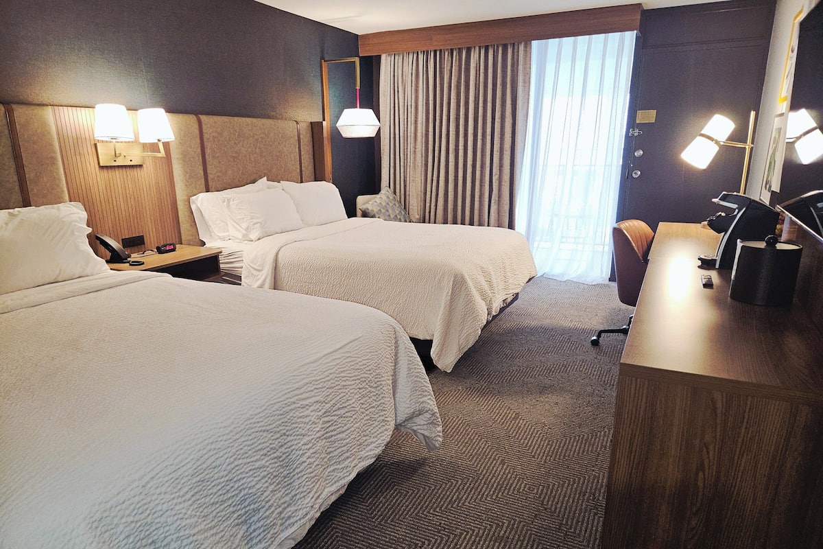 Habitación de hotel con dos camas, un escritorio, una silla, una lámpara y un ventanal con cortinas.