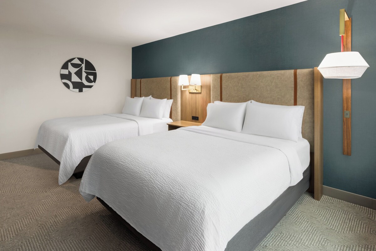 A hotel room with two beds, a wall clock, and a lamp.
