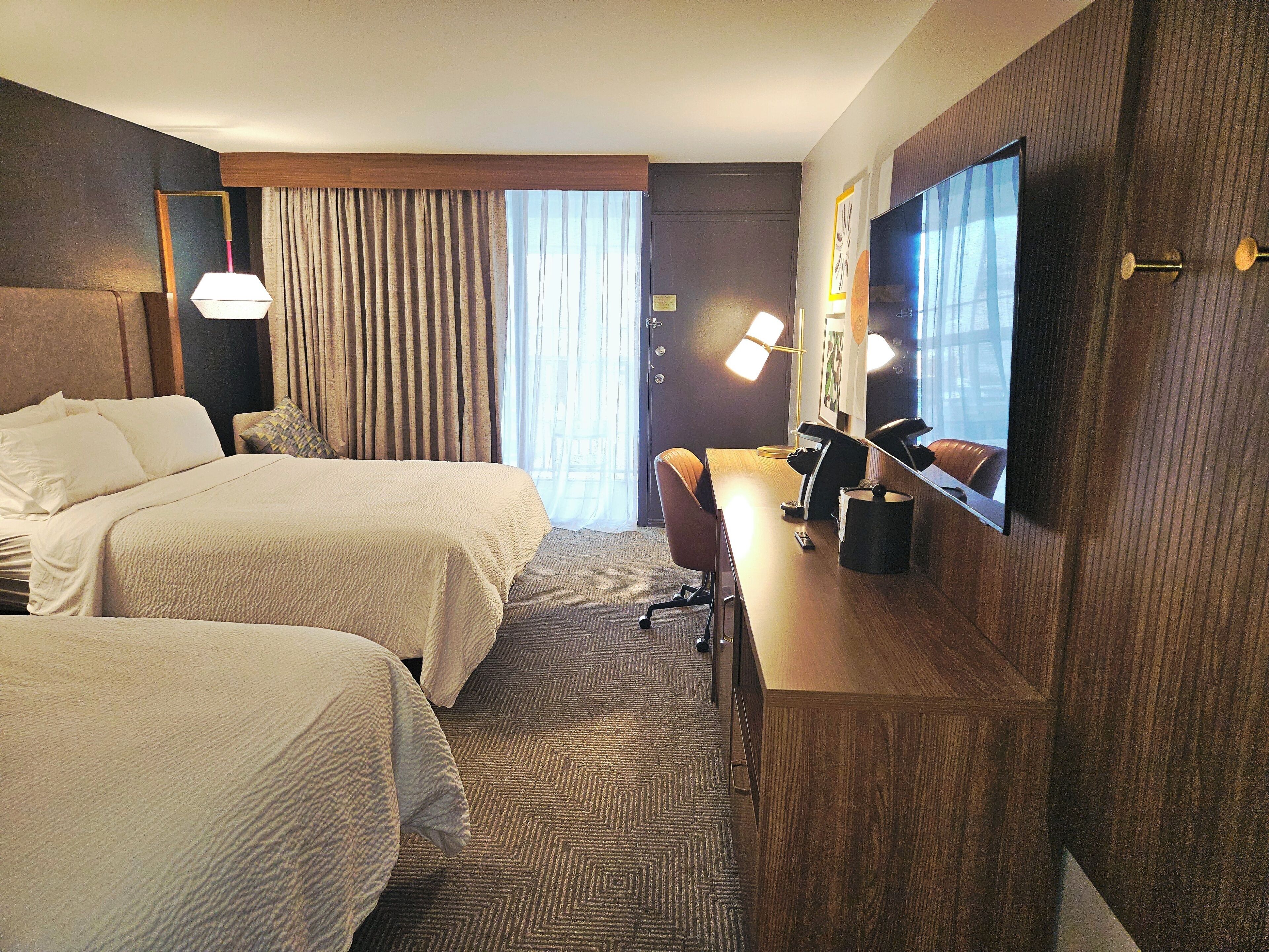 A hotel room with two beds, a desk, a chair, a TV, and a window with curtains.
