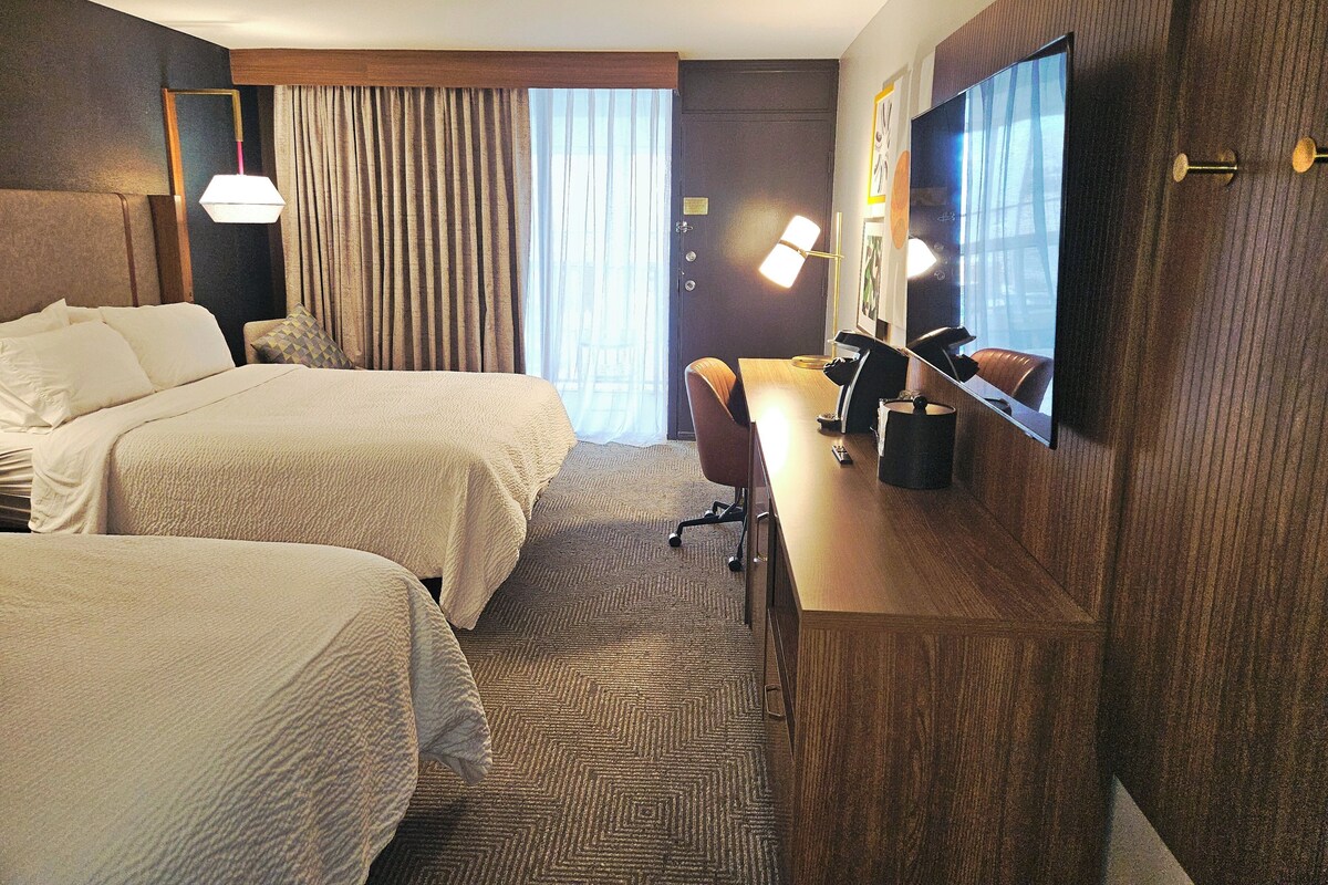 Habitación de hotel con dos camas, un escritorio, una silla, un televisor y un ventanal con cortinas.