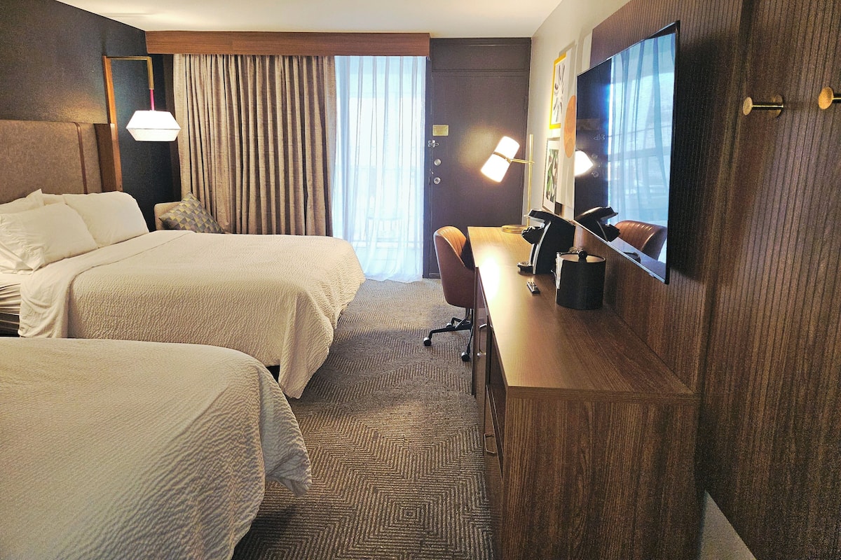 A hotel room with two beds, a desk, a chair, a TV, and a window with curtains.