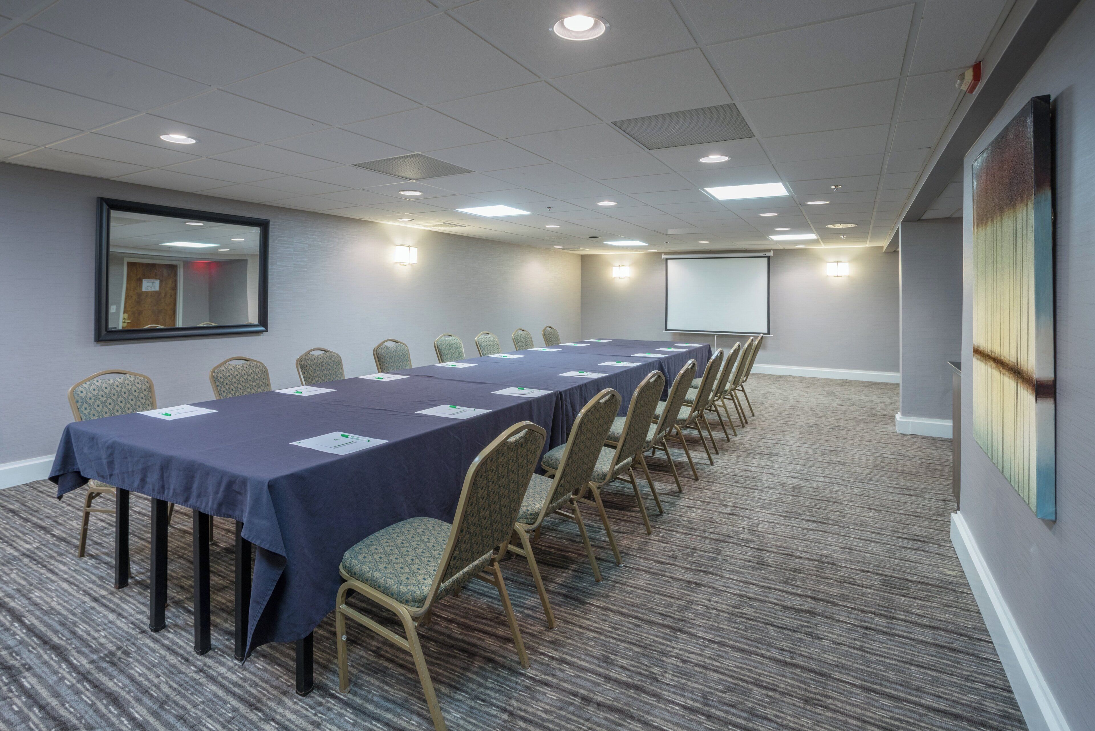 A conference room with a long table, chairs, a projector screen, and a large mirror on the wall.