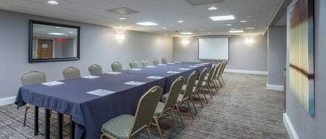 A conference room with a long table, chairs, a projector screen, and a large mirror on the wall.
