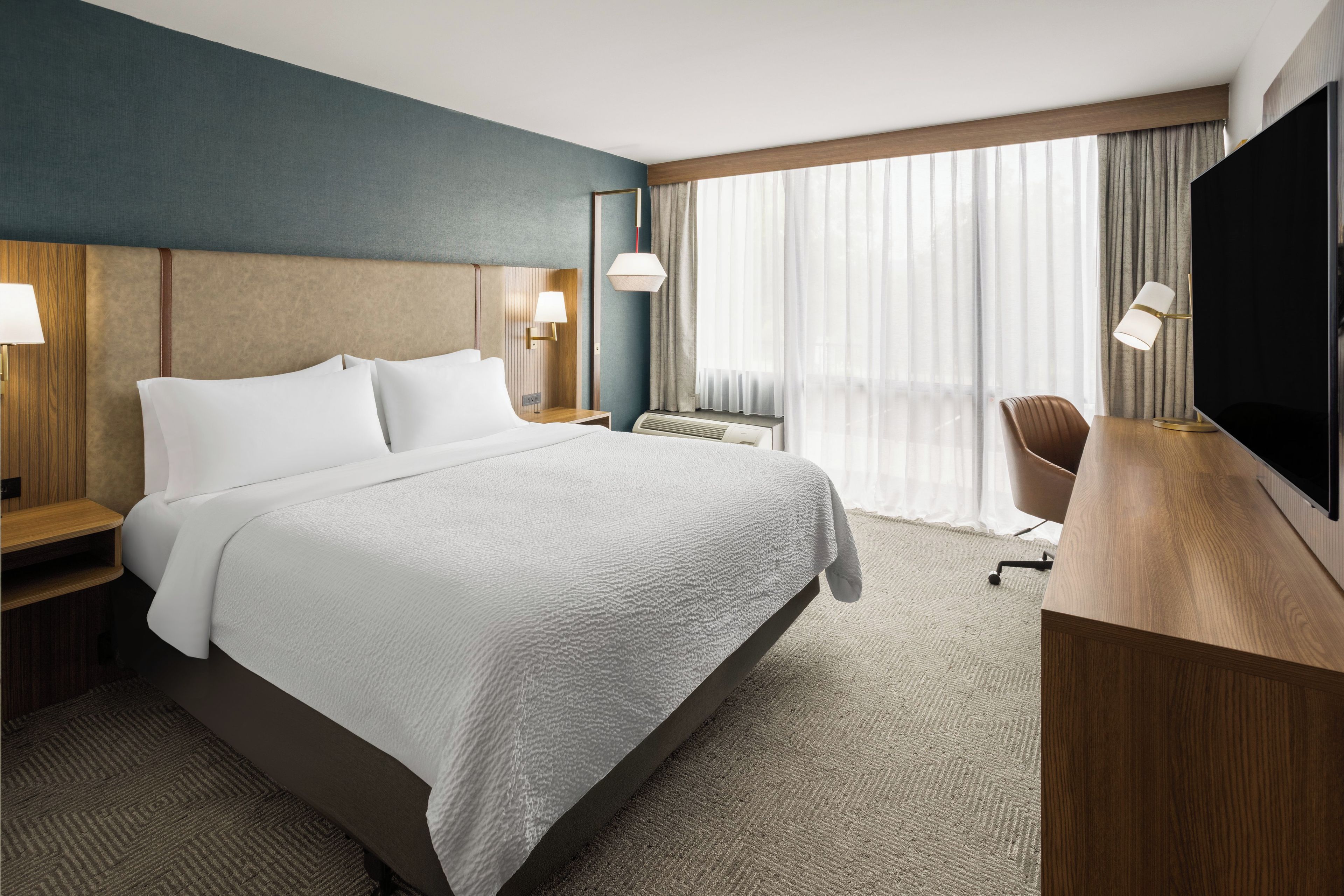 Habitación de hotel con una cama grande, mesitas de noche con lámparas, un escritorio con silla, un televisor y un ventanal con cortinas.
