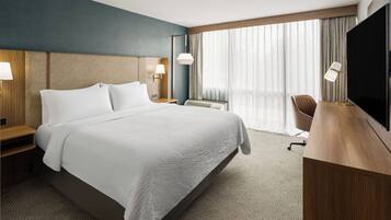 Habitación de hotel con una cama grande, mesitas de noche con lámparas, un escritorio con silla, un televisor y un ventanal con cortinas.
