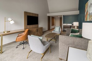 A hotel room with a flat-screen TV mounted on the wall, a desk with a lamp, a brown leather chair, a grey armchair, a patterned sofa, and a small round table. - Holiday Inn Harrisburg - Hershey Area, I-81 by IHG (Grantville)