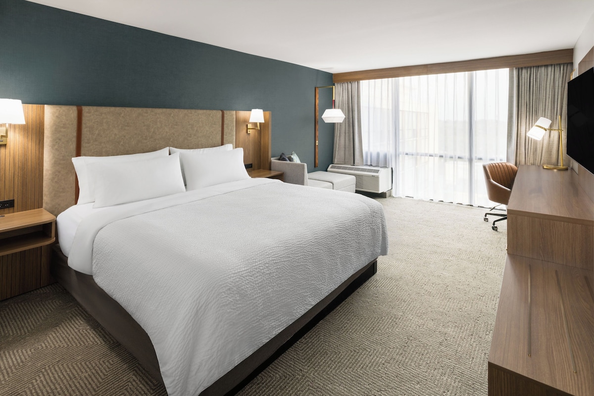 A hotel room with a large bed, bedside tables, a desk, a chair, a TV, and a window with curtains.