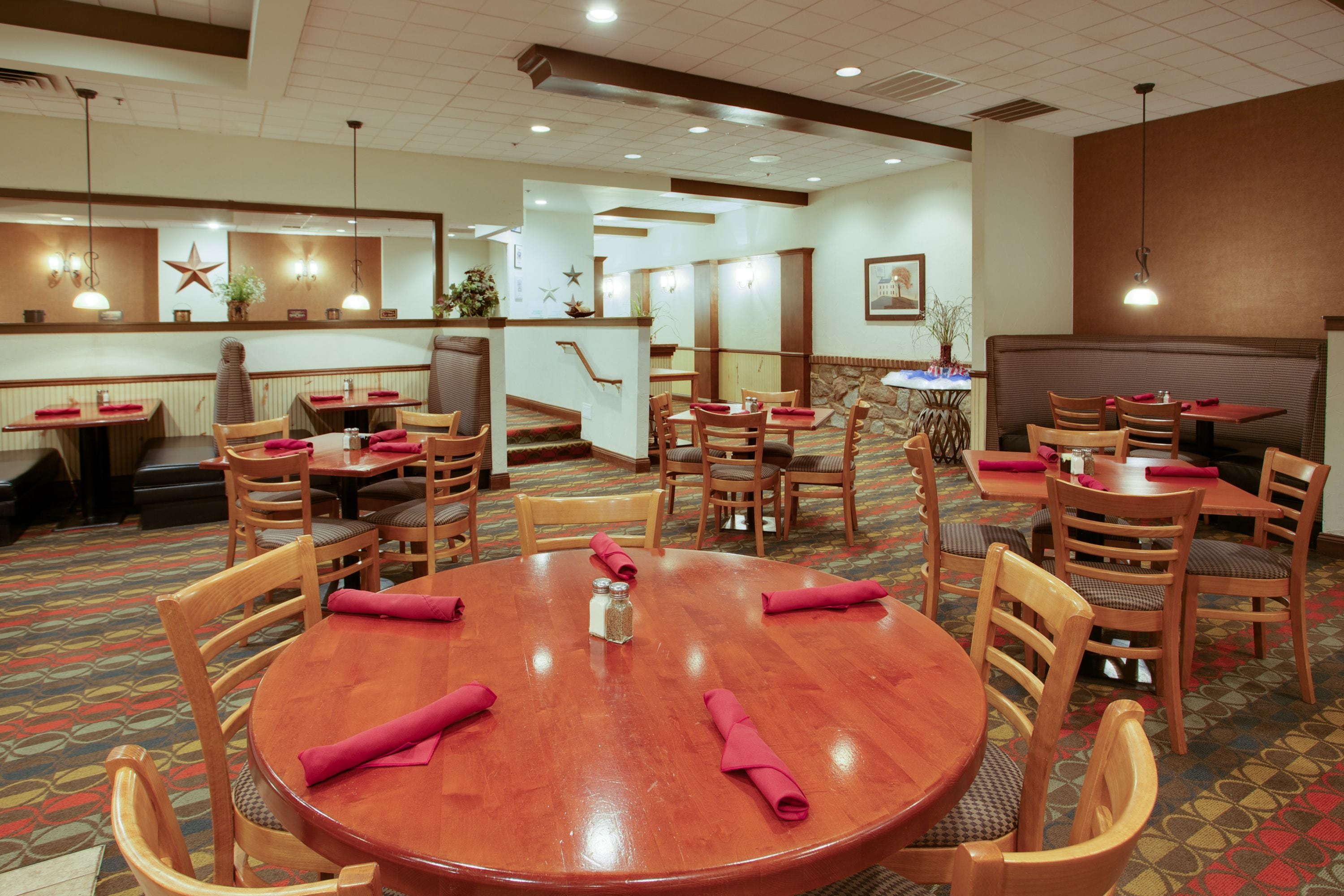 Zona de comedor de hotel con varias mesas redondas y sillas de madera, pisos alfombrados y una escalera que conduce a otro nivel.