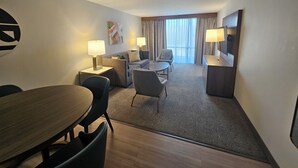 A hotel room with a dining table, two armchairs, a sofa, a TV, and a window with curtains.