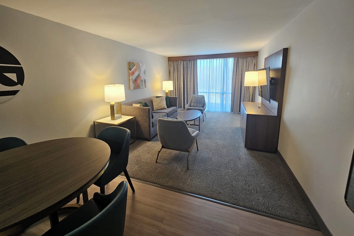 A hotel room with a dining table, two armchairs, a sofa, a TV, and a window with curtains.