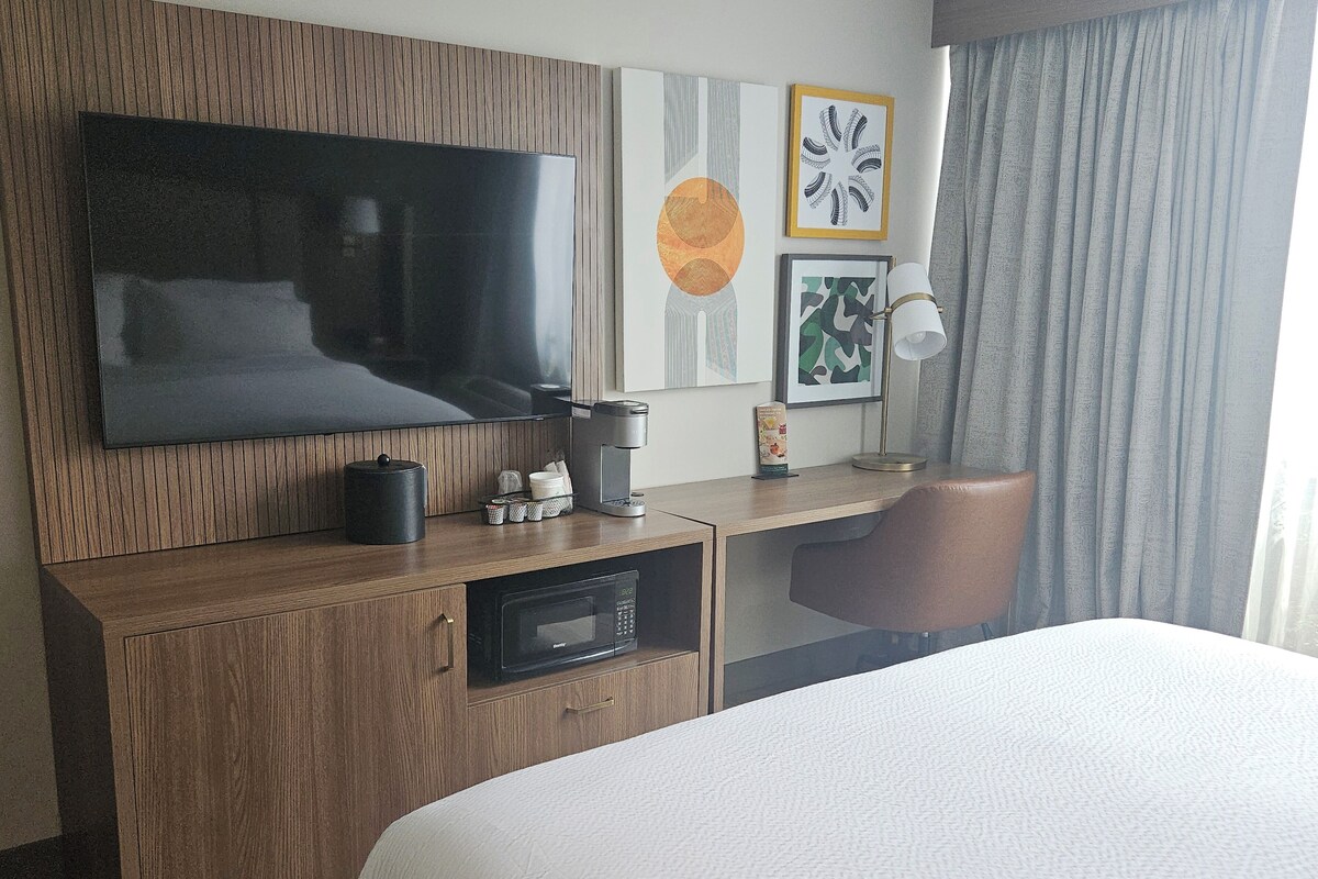 Habitación de hotel con una cama, televisor montado en un panel de madera, escritorio con cafetera, lámpara y obras de arte en la pared.