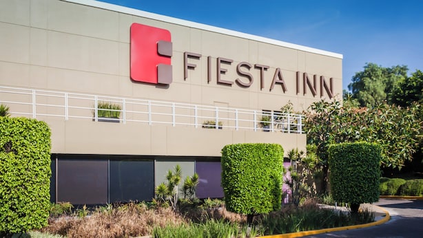 Edificio de Fiesta Inn con fachada moderna, ventanas grandes y jardines bien cuidados.