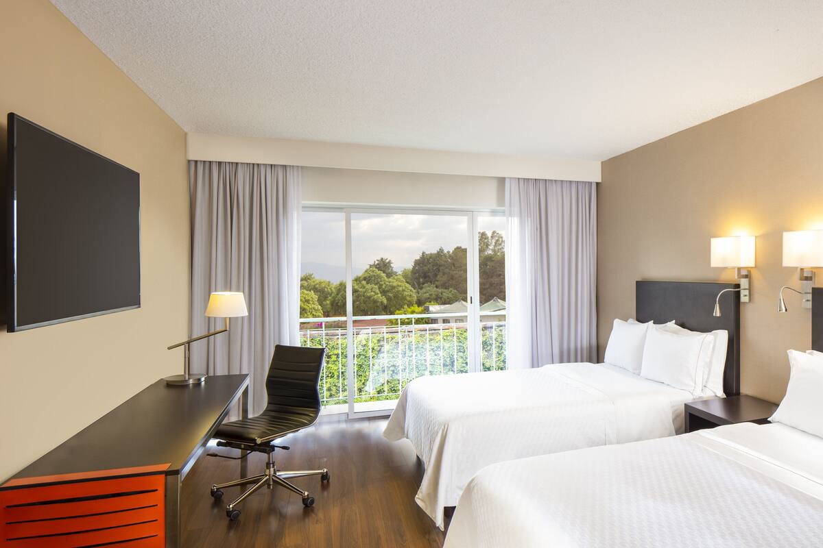 A hotel room with two beds, a desk, a chair, a TV, and a balcony with a view of greenery.