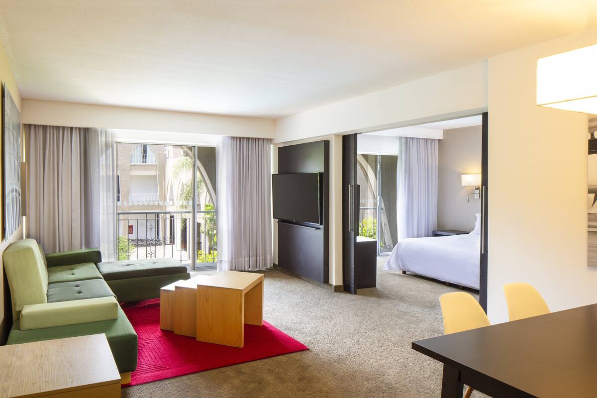 A hotel room with a green sofa, a wooden coffee table, a TV, a bed, and a balcony with curtains.
