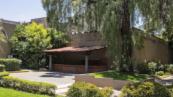 Un patio con zona de estar cubierta, rodeado de vegetación cuidada y un árbol grande.