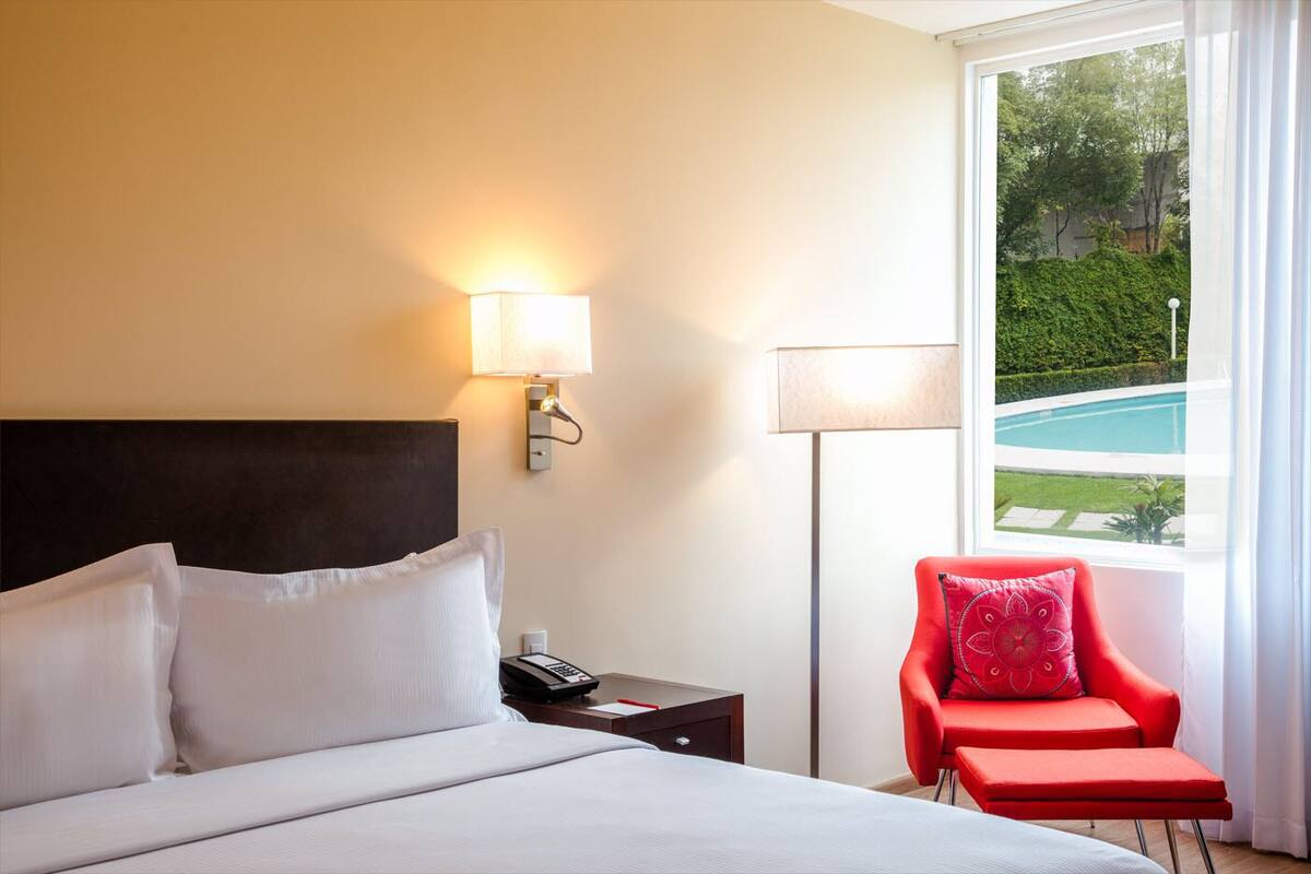 A hotel room with a bed, a nightstand with a phone, a red chair, and a view of a swimming pool through large windows.
