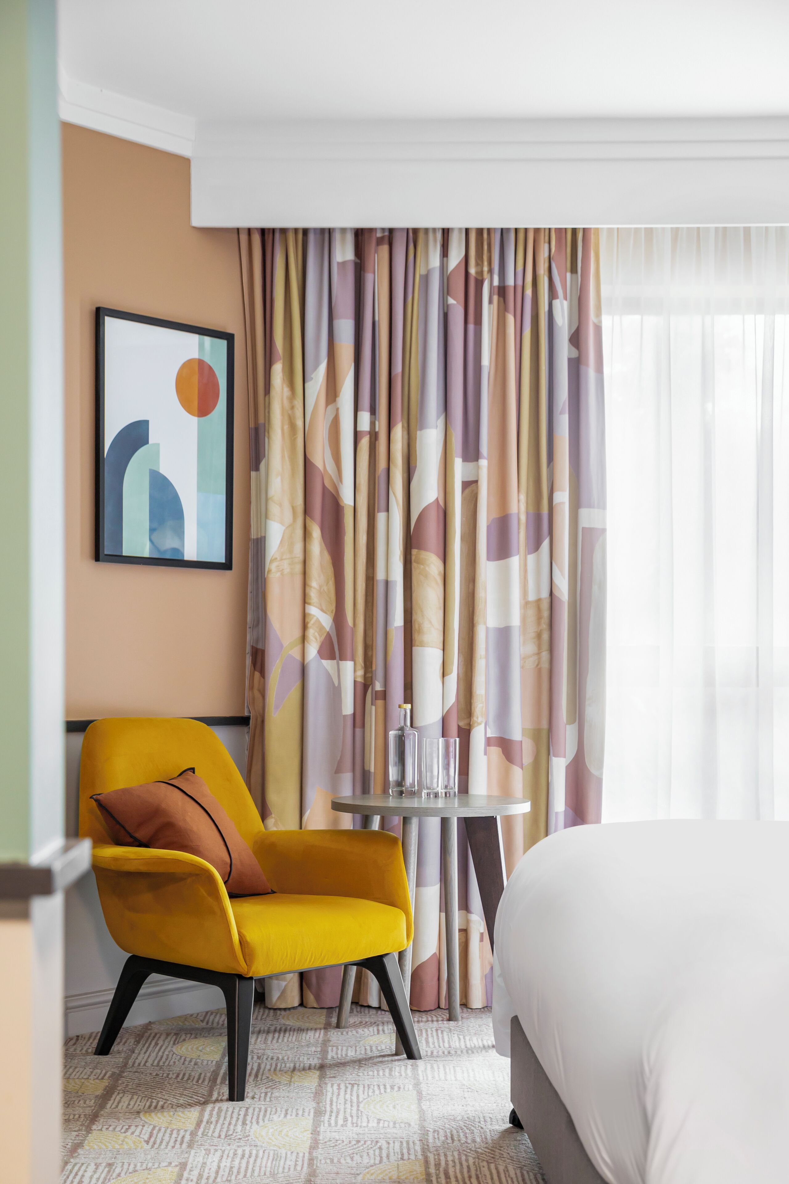 A hotel room with a bed, a yellow armchair, a small table with a glass, and a curtain with a geometric pattern.