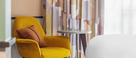 Habitación de hotel con cama, un sillón amarillo, una mesita con un vaso y una cortina con diseño geométrico.