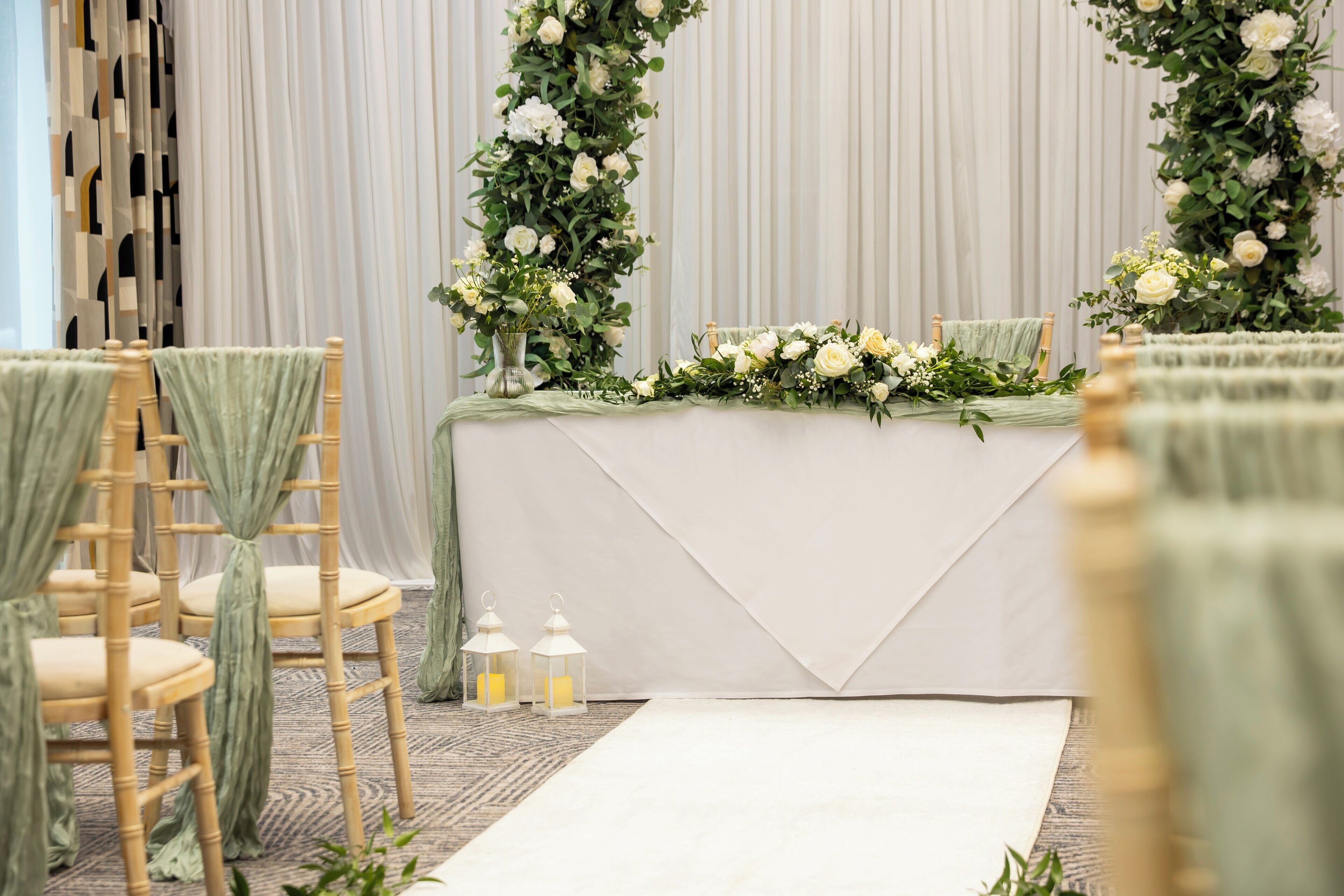 A wedding ceremony setup with a white table, floral arrangements, and chairs with green drapes.