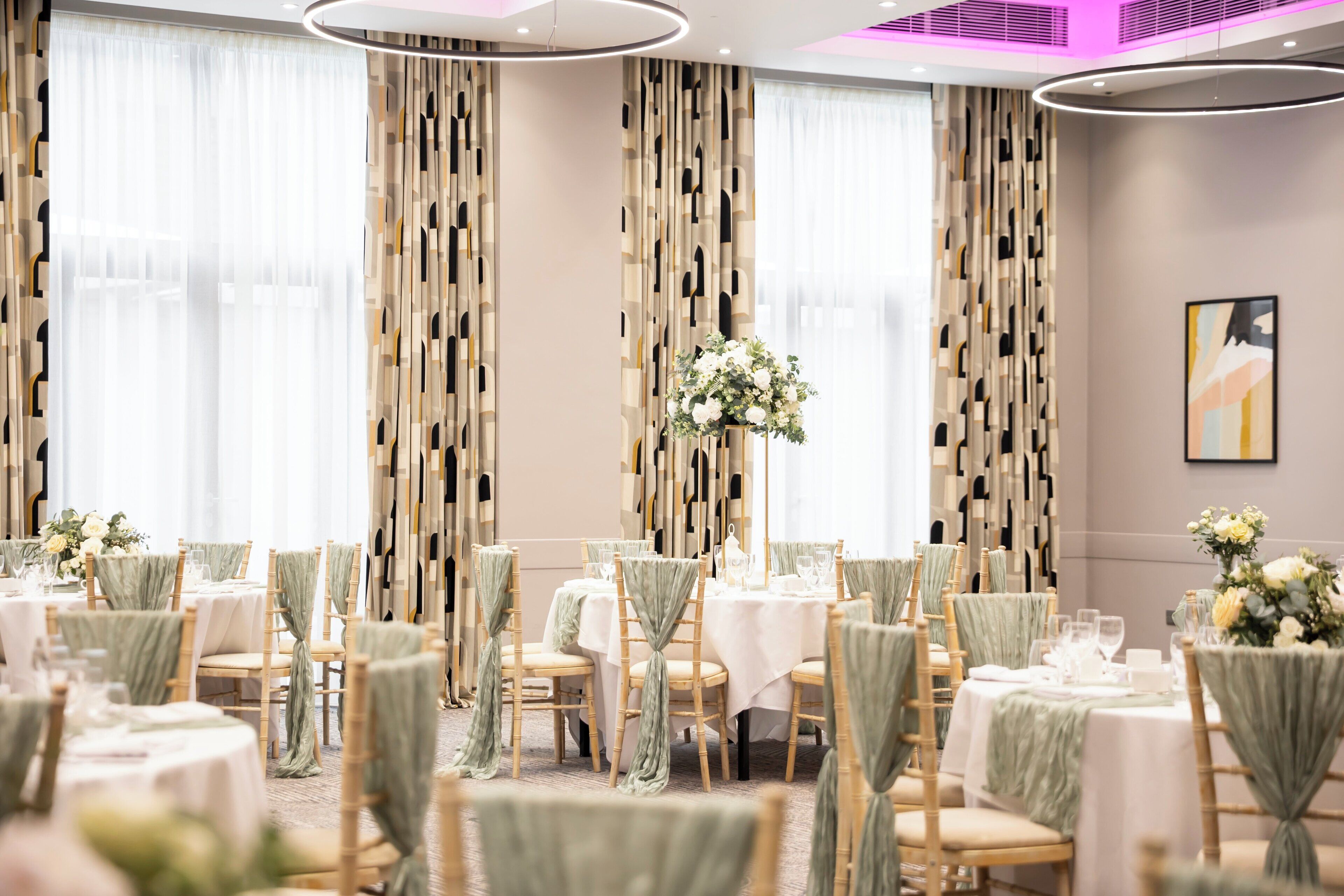 Un salón de banquetes con mesas redondas, sillas con cintas verdes y un centro de mesa floral.