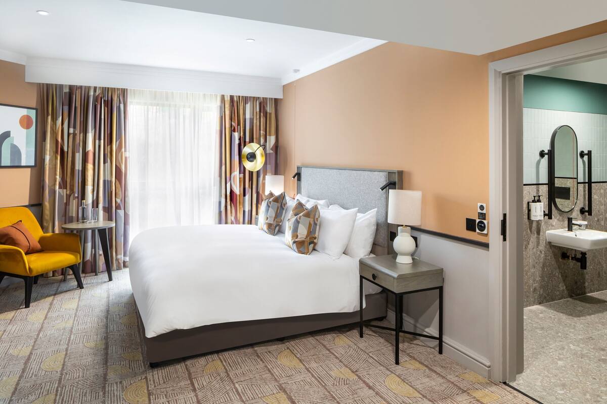 A hotel room with a large bed, a bedside table with a lamp, a small round table, a yellow armchair, and a view of the bathroom through an open door.