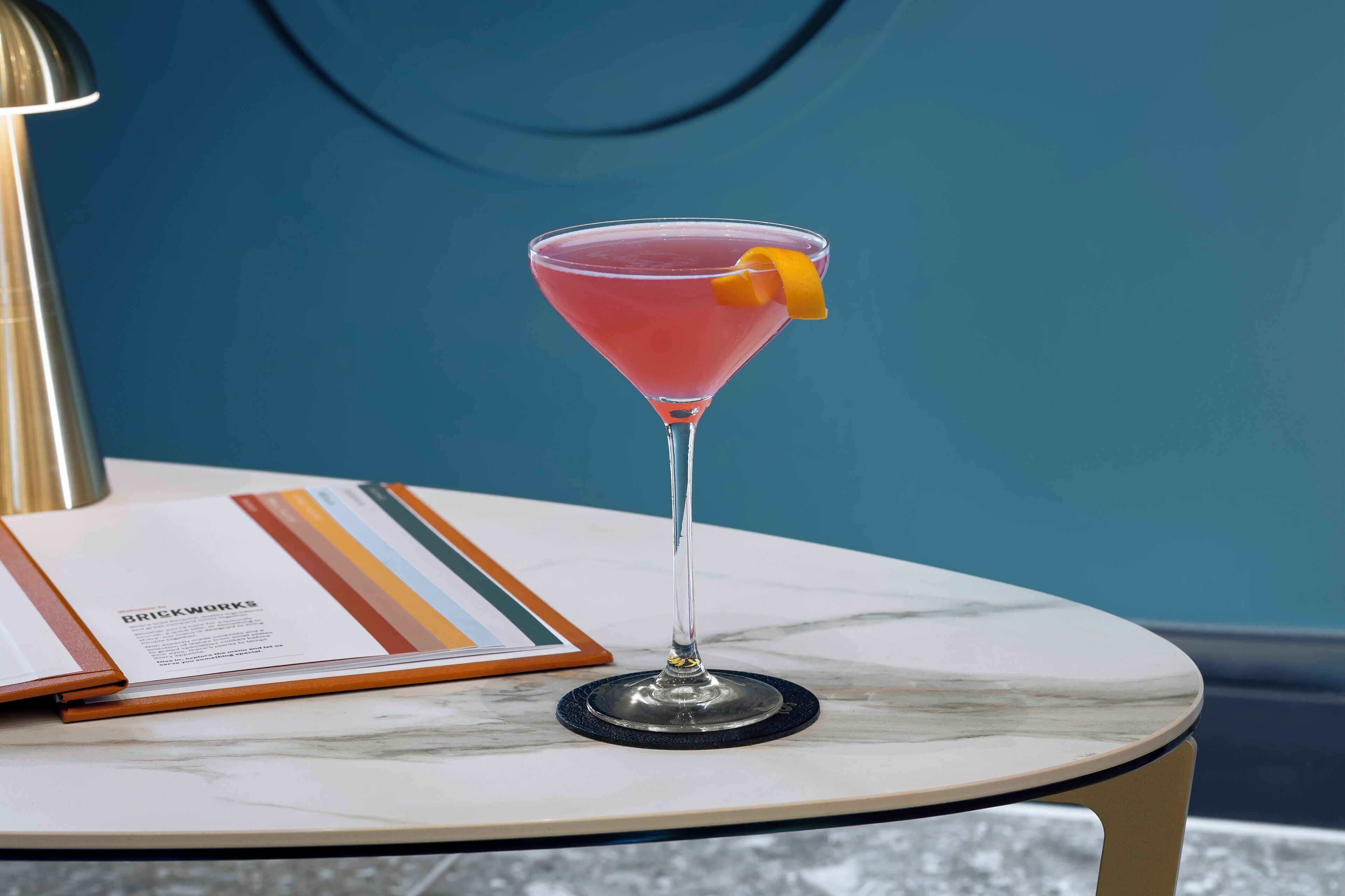 A martini glass with a pink drink and an orange peel garnish on a marble table.
