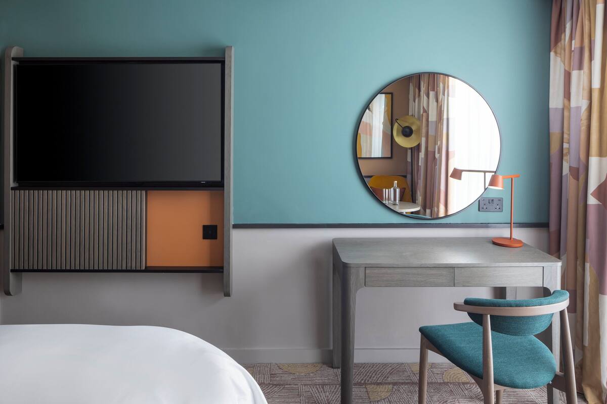 A hotel room with a bed, a desk with a chair, a TV, a round mirror, and a lamp.