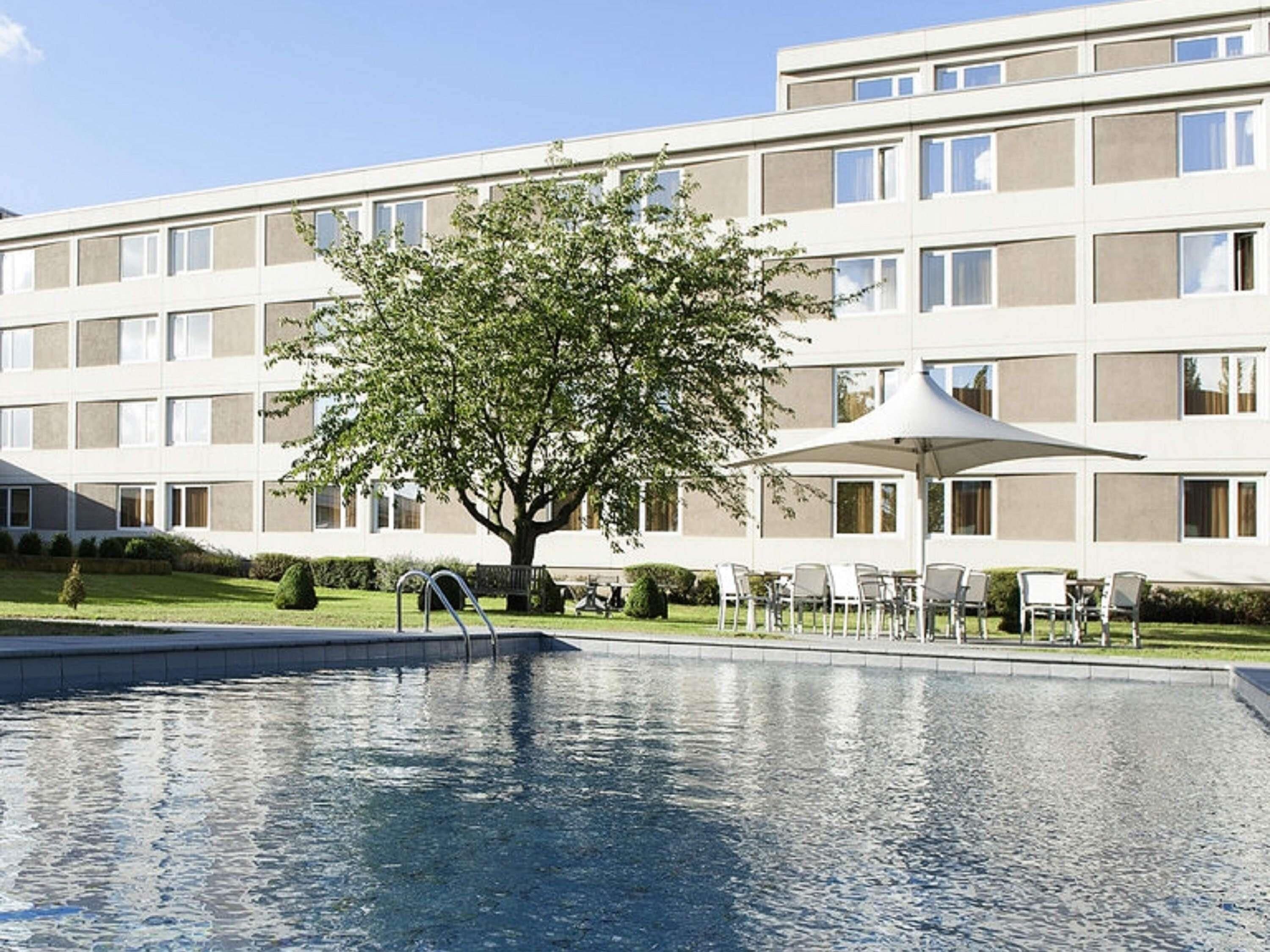 A modern hotel with a swimming pool, outdoor seating, and a large tree in the center.