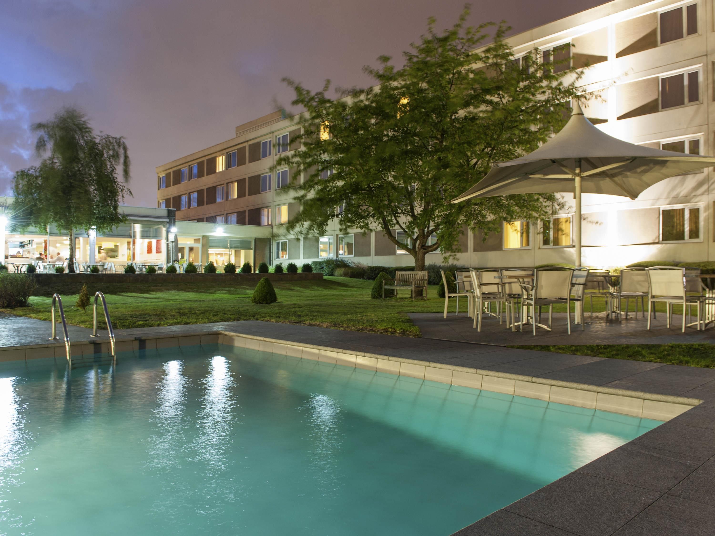 Uma piscina com um terraço ao redor, um edifício com várias janelas e uma árvore.