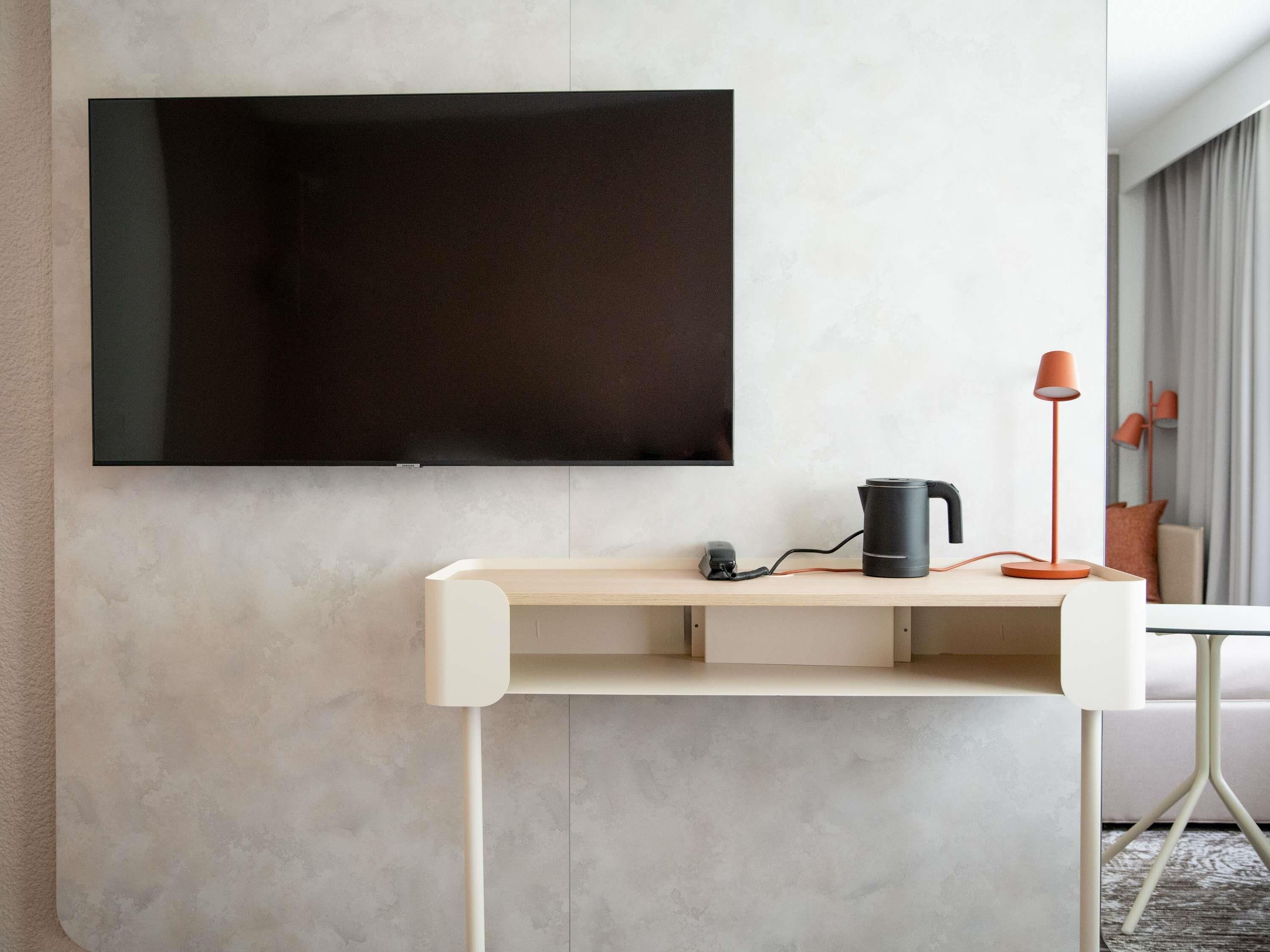 A modern room with a flat-screen TV mounted on the wall, a wooden desk with a kettle and lamp, and a white chair.