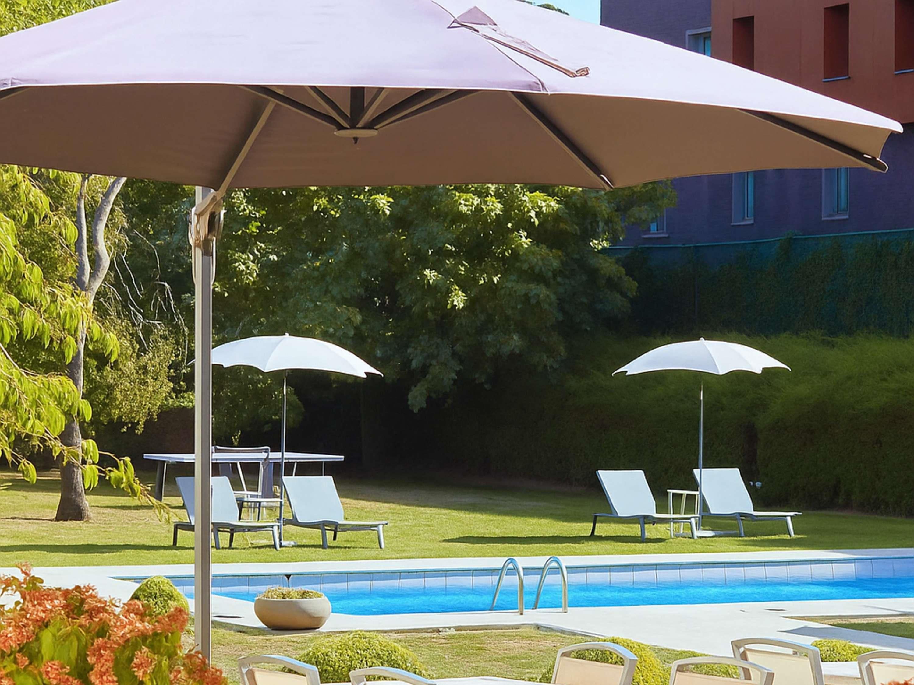 Ein Poolbereich mit zwei Liegestühlen und einem Tisch unter einem großen Sonnenschirm, umgeben von Grünpflanzen und einem Gebäude im Hintergrund.