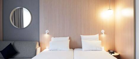 Camera d'albergo con due letti, un divano grigio, uno specchio rotondo e lampade da comodino.