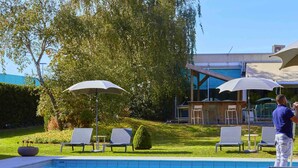 Una piscina con sillones de playa y sombrillas, rodeada de un césped cuidado y un edificio moderno al fondo.