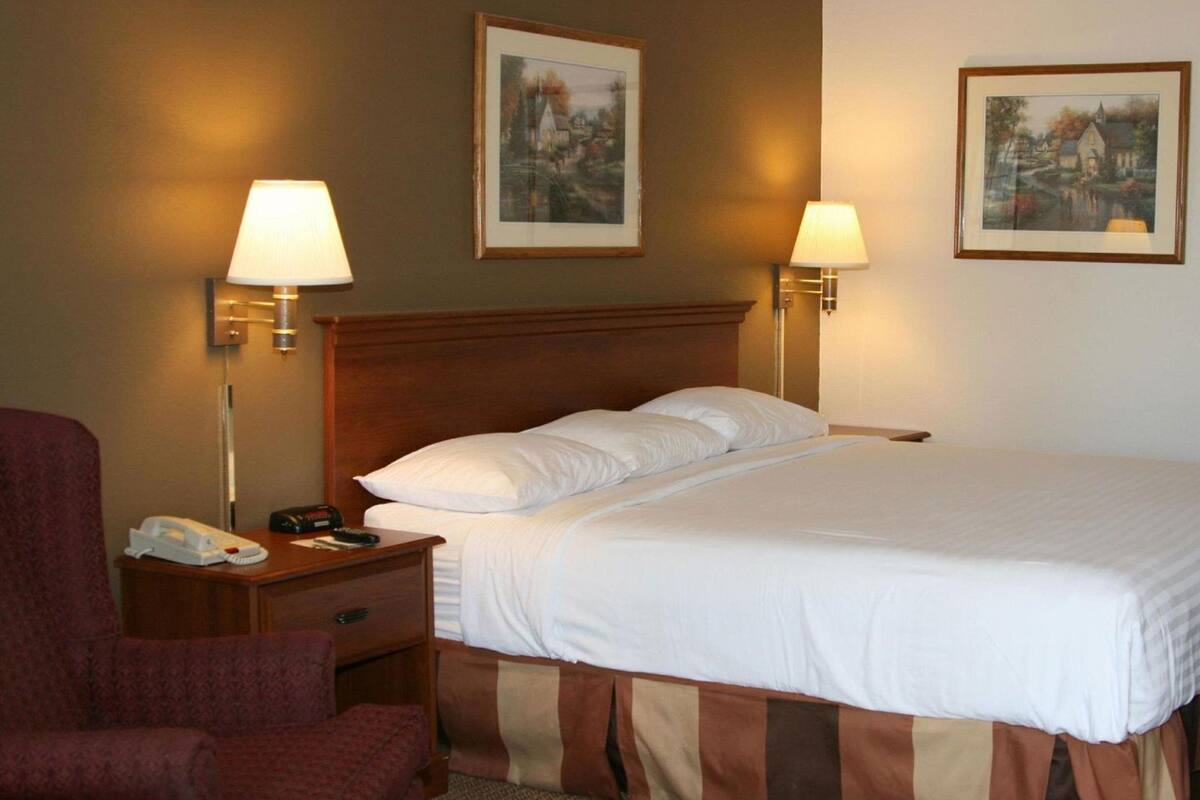 A hotel room with a bed, two bedside lamps, a chair, a nightstand with a phone and remote, and two framed pictures on the wall.