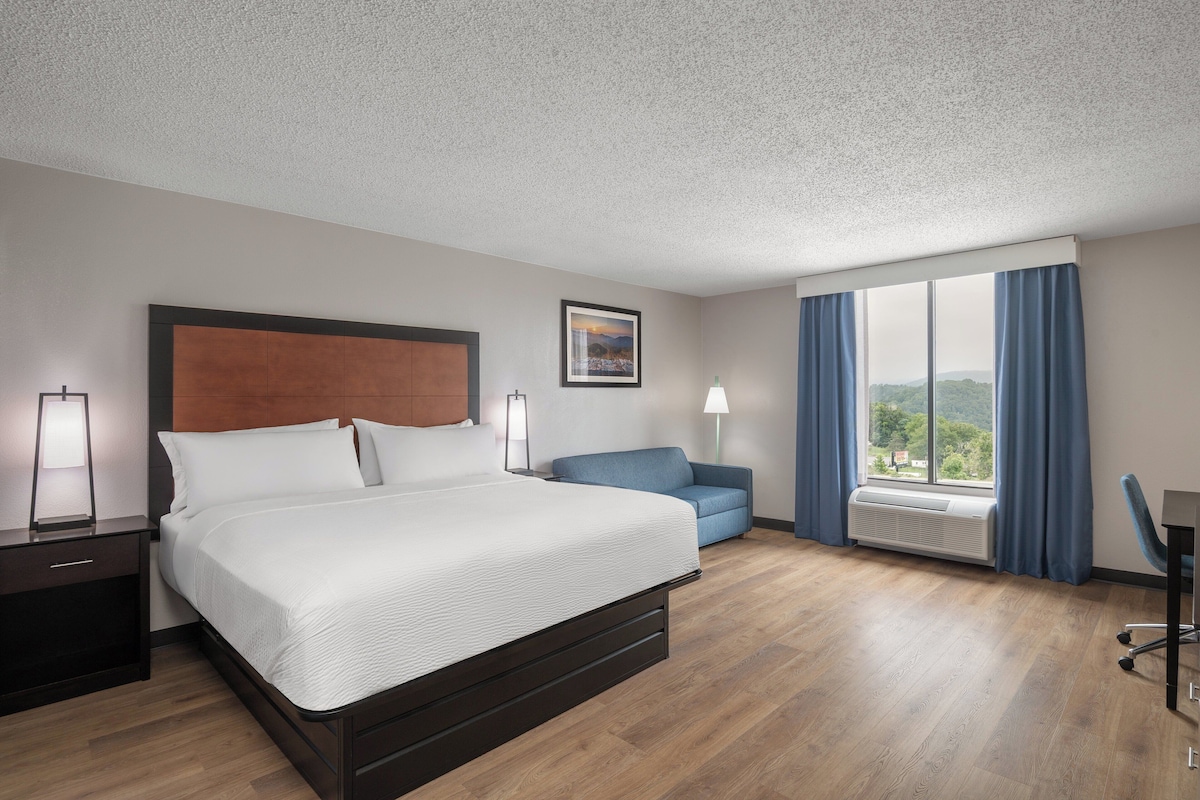 A hotel room with a bed, a desk, a chair, a window with curtains, and a wall-mounted air conditioner.